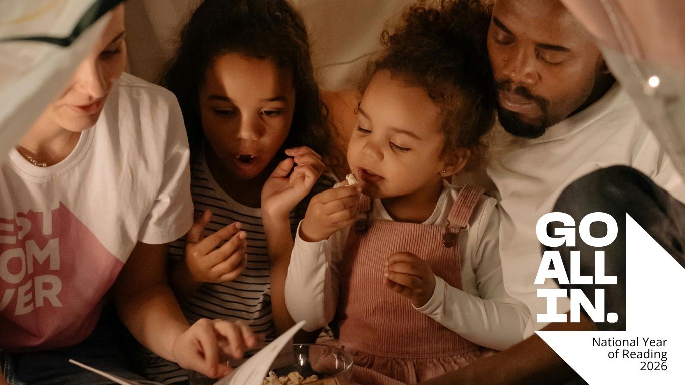 A young mum and dad sit under a white indoor tent festooned with lights with their two young children between them, as they eat popcorn reading a book