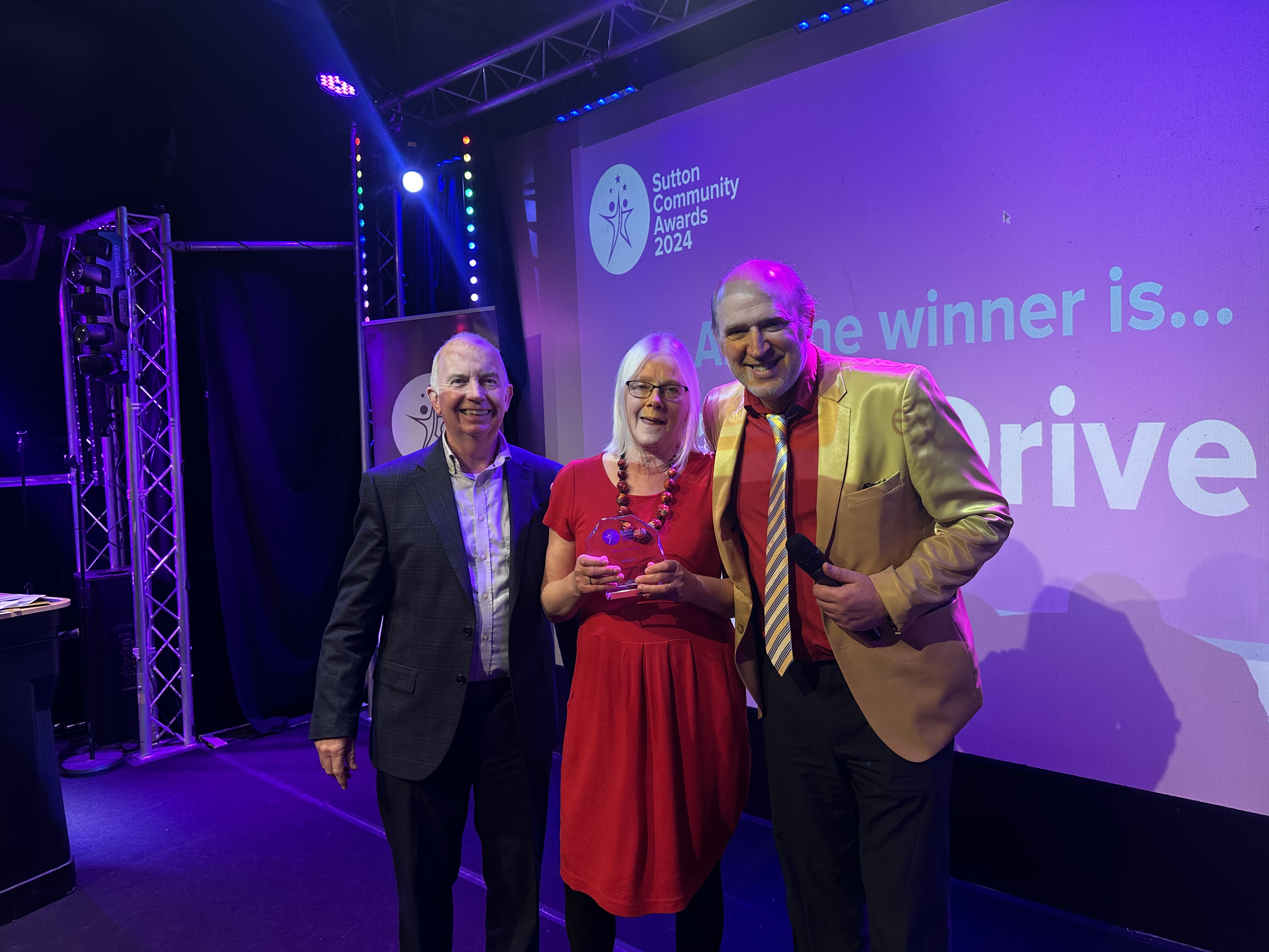 Leader of the Council Barry Lewis in a suit, Carol Driver wearing a red dress, and comedian Tim Vine wearing a gold jacket with a stripy tie and red shirt on stage at the Sutton Community Awards 2024