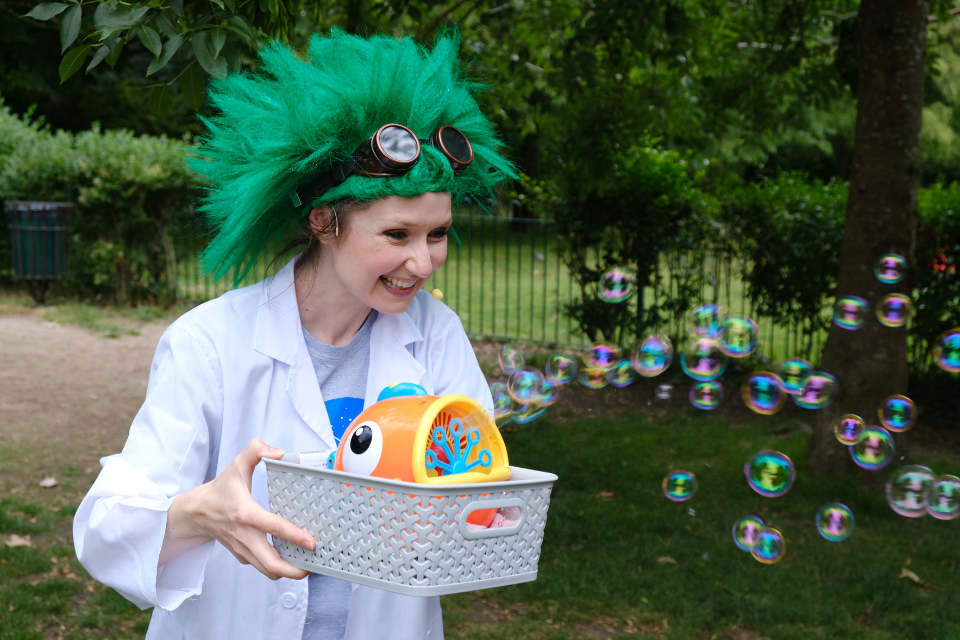 A young woman dressed in a lab coat and large green hair holding a bubble blowing machine