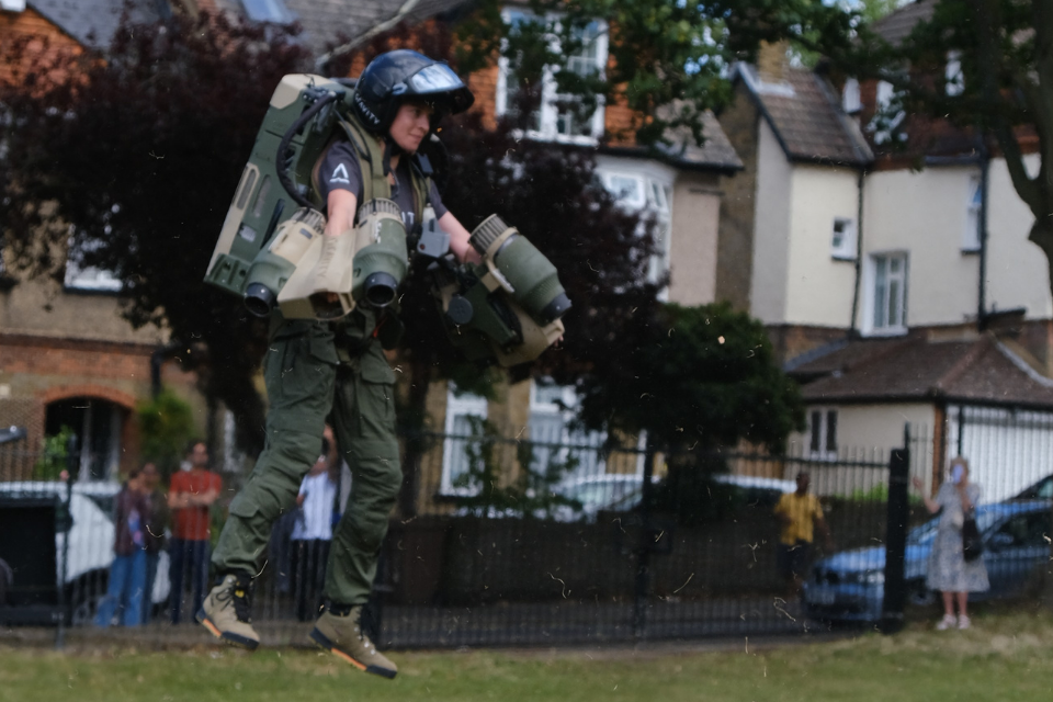 A woman hovering over the ground in a green and brown coloured flight suit with air thrusters attached to her arms