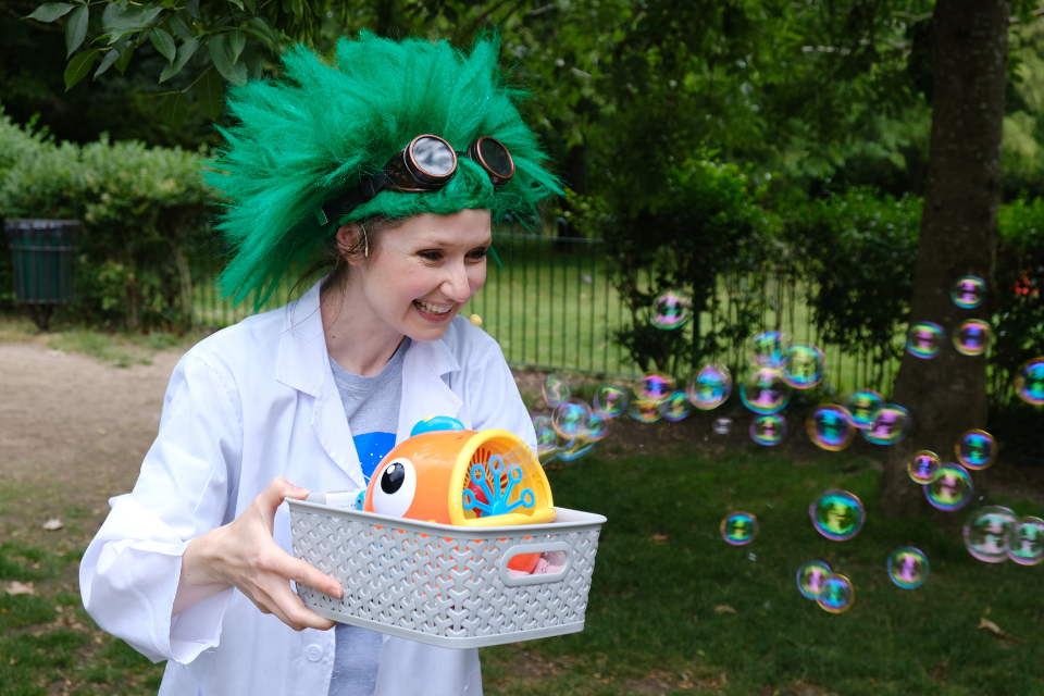A woman wearing a lab coat and large green wig operating a small toy automatically producing several bubbles