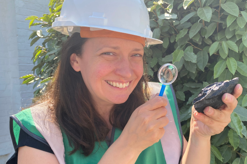 A smiling woman wearing a safety helmet and high vis jacket holding a magnifying glass and a piece of flint rock