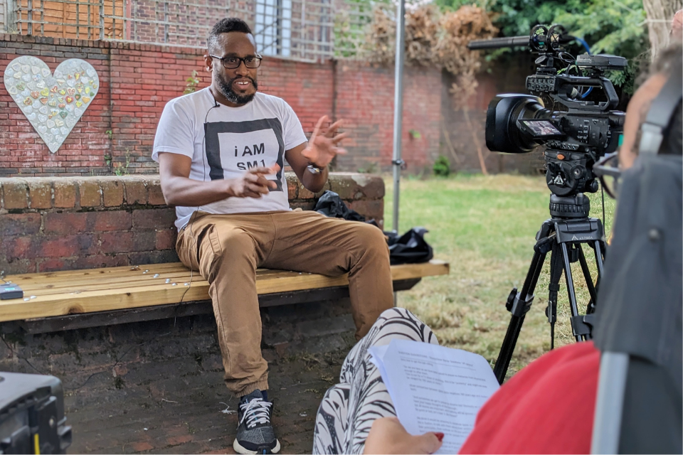 A man sat on a bench outside speaking towards a film camera