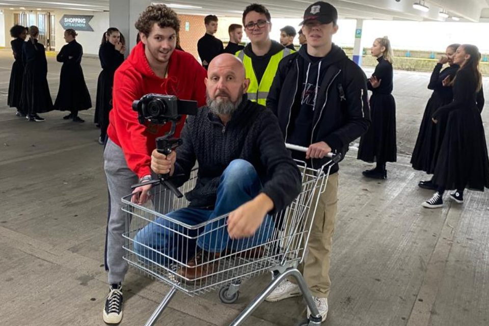 A man crouched in a shopping trolley while holding a camera and being pushed by three young men