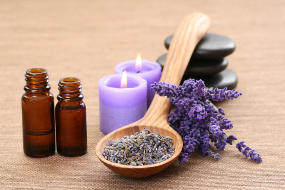 Lavender Herbs on a wooden spoon next to two small brown bottles and two lit purple candles