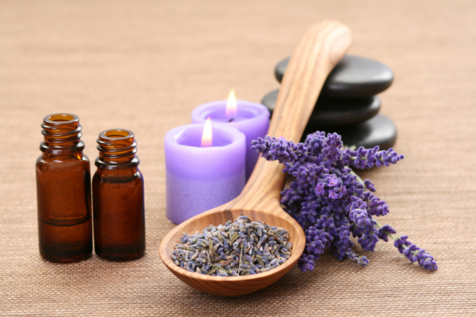 A wooden spoon holding lavender herbs, next to two lit lavender candles, two small jars of oil and a small number of sprigs of lavender