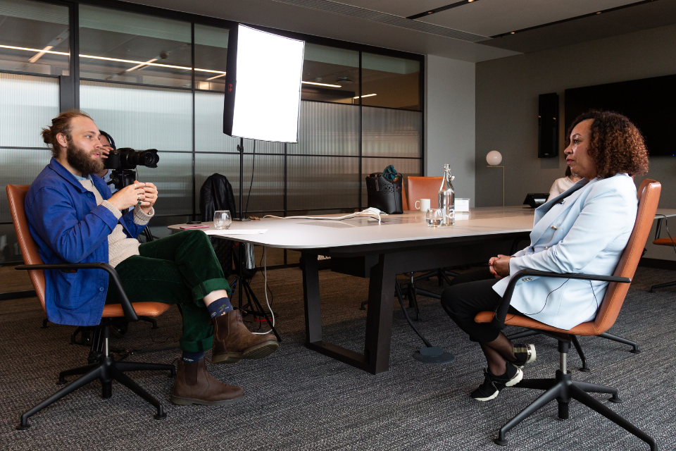 An interview between a man and a woman sat opposite each other across the corner of a table with a camera placed in the background recording the two of them speaking