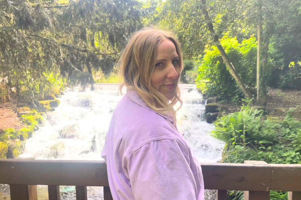 A smiling young blonde woman stood on a bridge overlooking a small waterfall