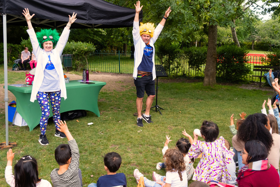 Two adults wearing bright coloured wigs with their hands extended in the air
