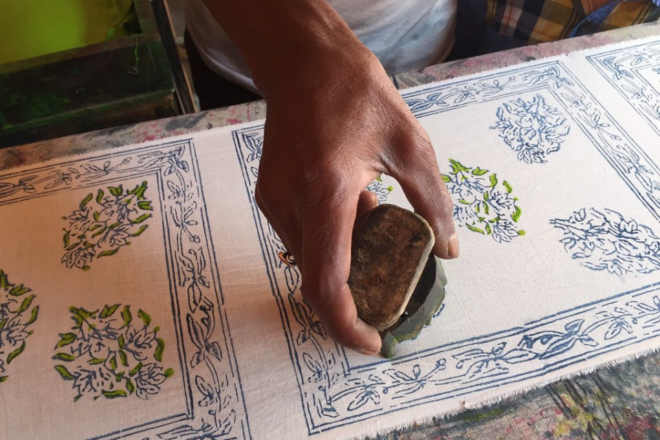 A hand pressing a block of wood that has been dipped in dye onto a cream-coloured cloth. The cloth has several leaf patterns already printed onto it.
